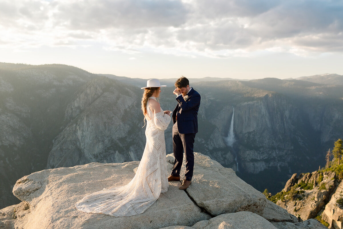 A groom wipes his eyes while exchanging vows with his bride while atop Taft Point with the view of Yosemite falls in the background at Yosemite National Park. 