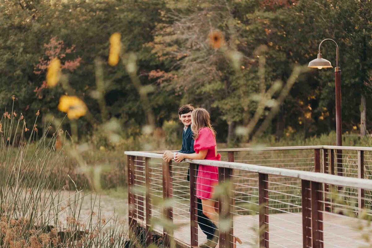 far back view of engaged couple, talking while standing on a bridge, with florals in the foreground.