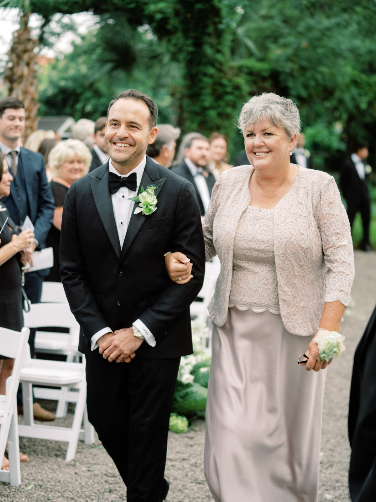 A joyous moment at a wedding ceremony, featuring a groom happily walking down the aisle with his mother, showcasing love and tradition in a beautiful outdoor setting.