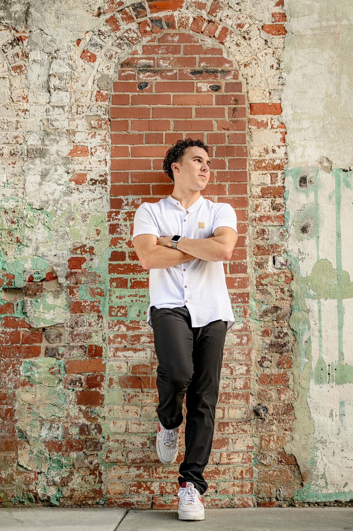 Senior portrait of a boy wearing dark pants and white shirt leaning against a teal and red brick wall for his graduation portraits