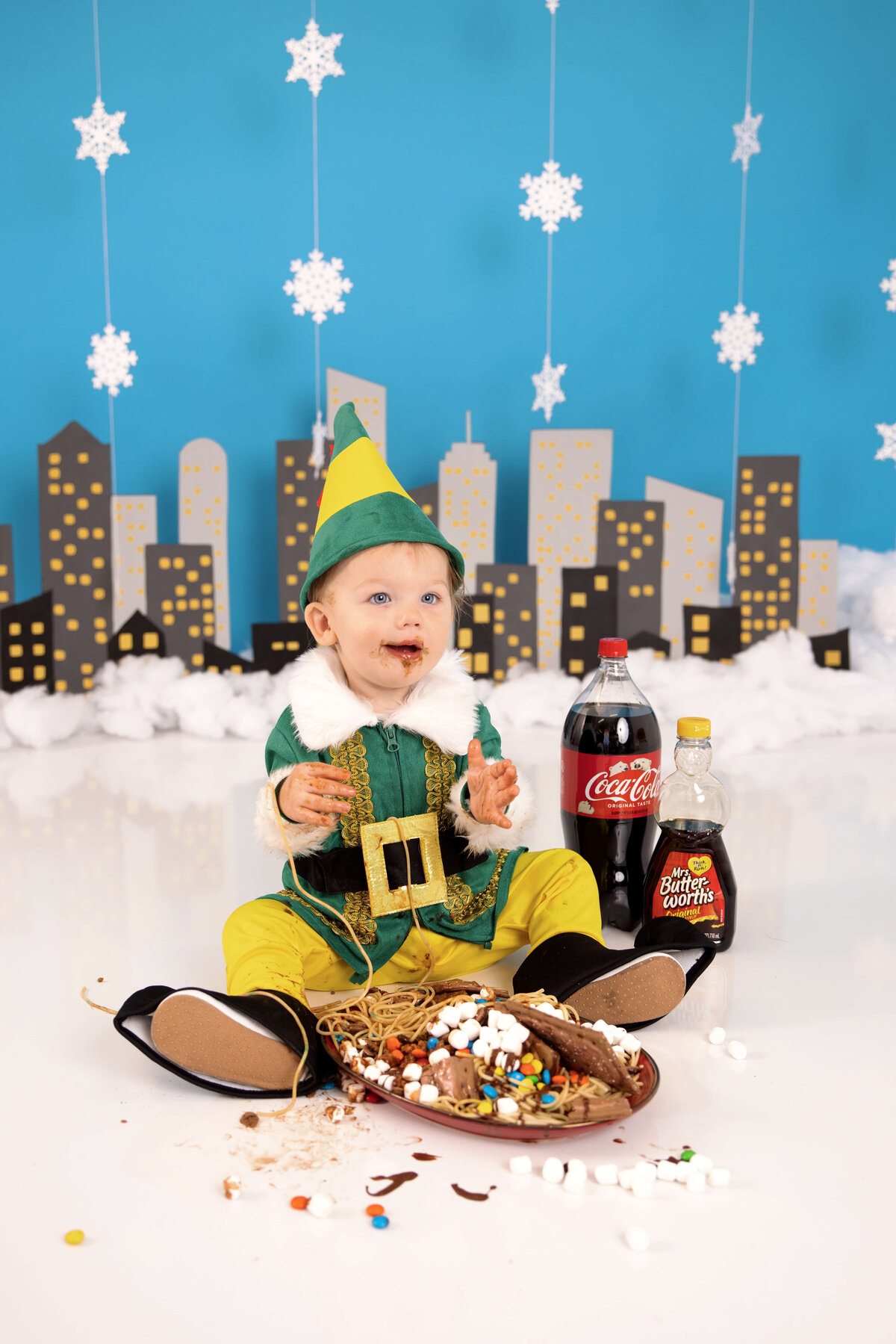 A baby in a green elf costume sits on a white floor, surrounded by chocolate syrup and candy, captured beautifully by a Jacksonville cake smash photographer. Nearby are a Coca-Cola bottle and a syrup bottle. The background shows a cityscape silhouette with hanging snowflakes.