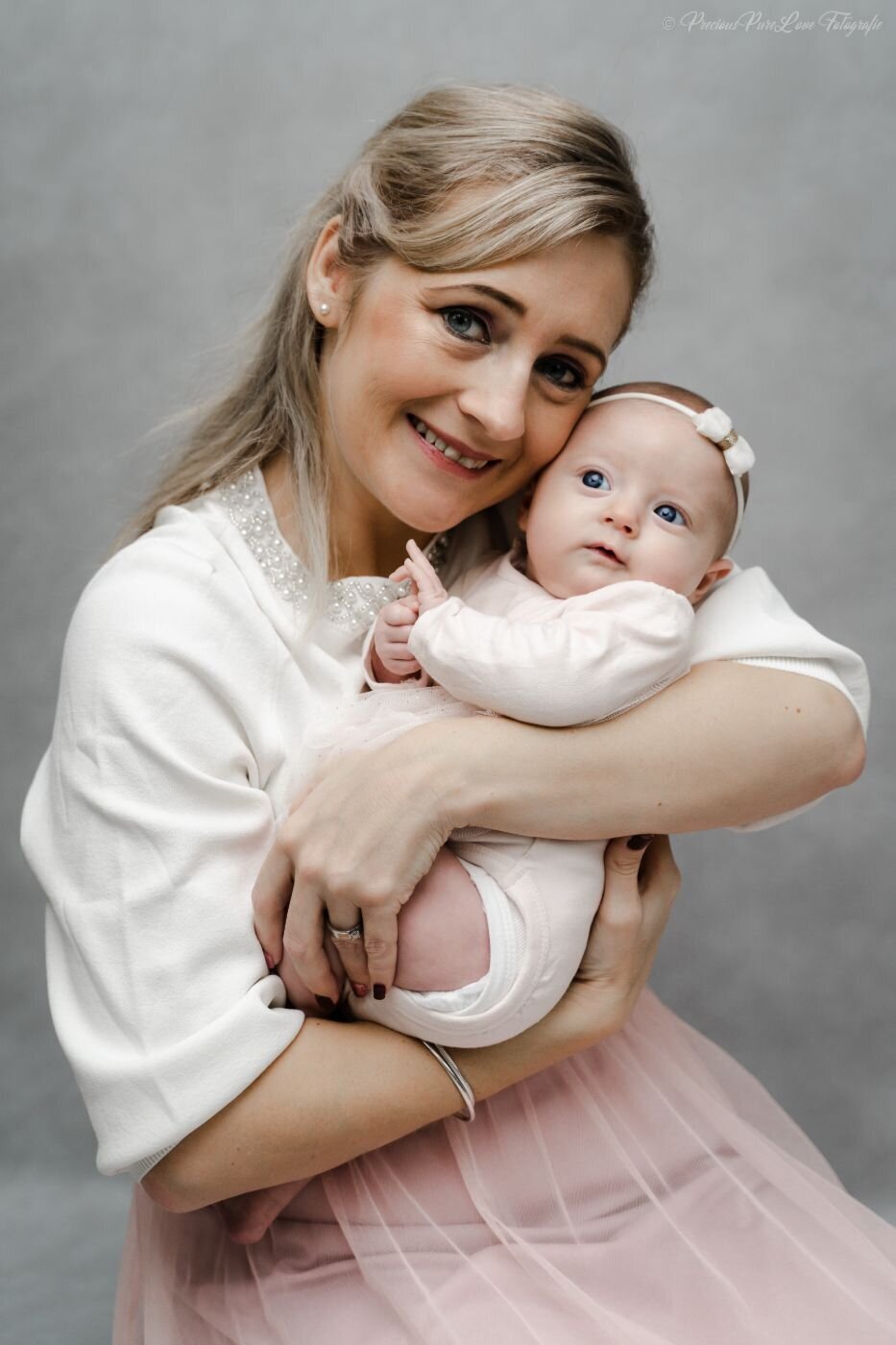 A candid, color portrait of a mother holding her newborn baby close to her chest. The mother, with a warm smile, looks directly at the camera. The baby, wearing a white outfit and a small white bow, is looking off to the side. The mother wears a white sweater with a beaded collar and a pink tulle skirt.