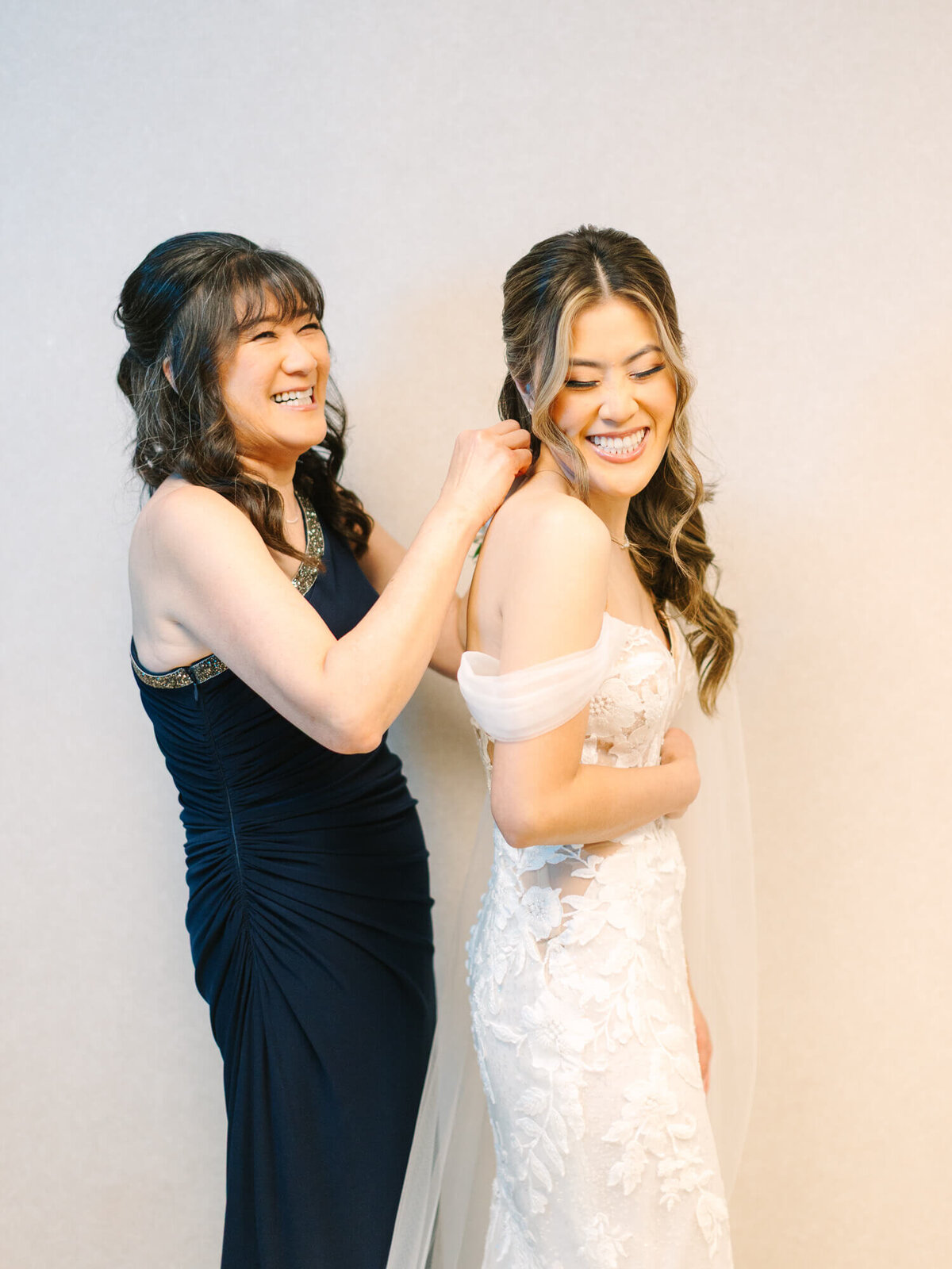 A smiling bride in a white lace gown is being helped with her dress by her mom in a navy dress. They share a joyful moment, conveying warmth and happiness.