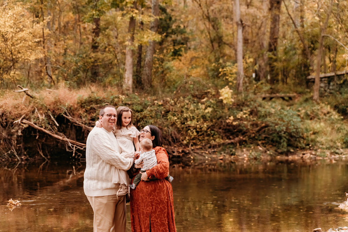 Mother and father holding their two young children in their arms in front of a creek.  Mom is wearing burnt orange dress from baltic born, and daughter is wearing ivory dress from Joyfolie.  Photo taken by Philadelphia Family Photographer, Kristi