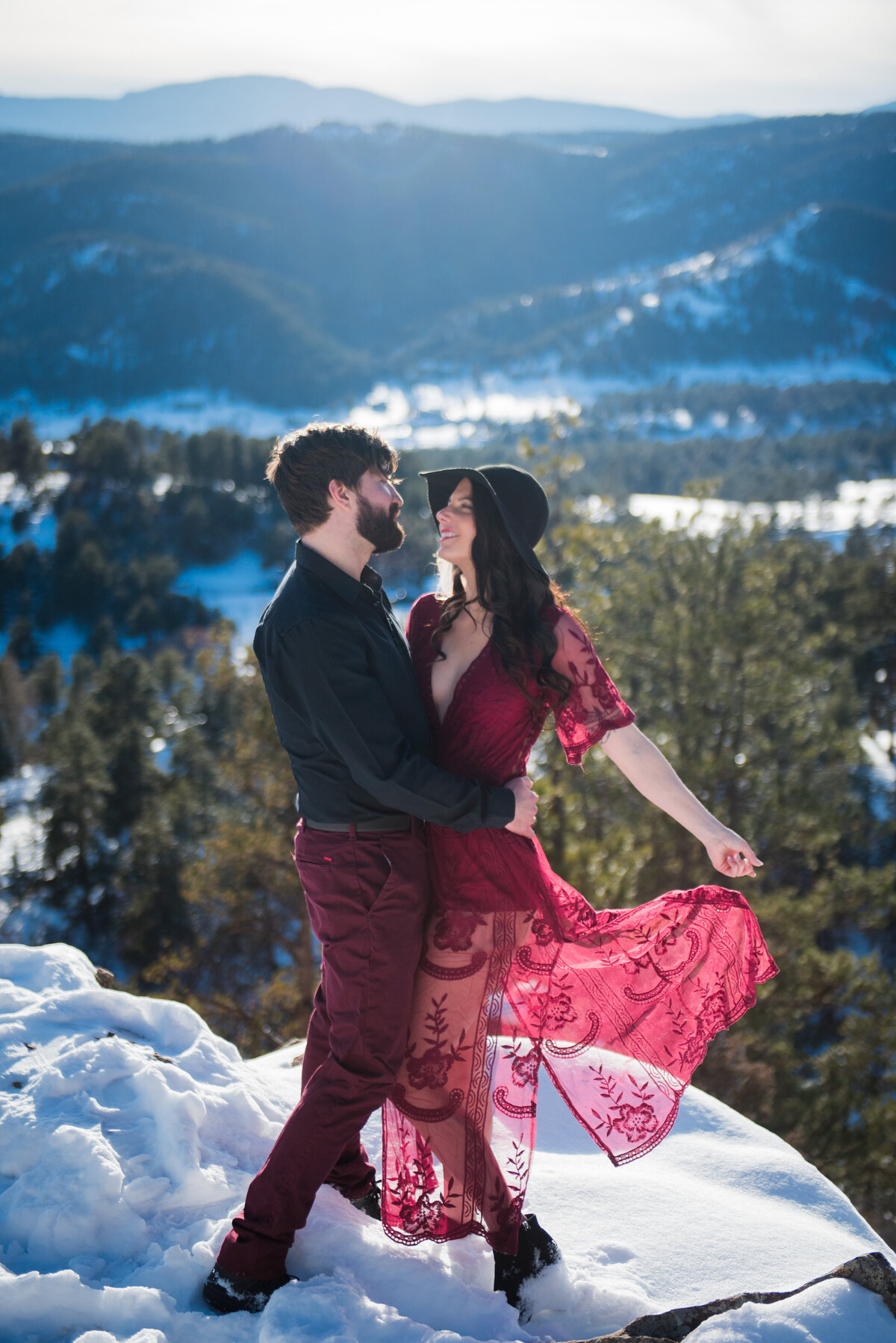 A man and woman standing on the top of Mount Falcon during their engagement session. The man has his hands around the woman's waist as she plays with the train of her red dress.