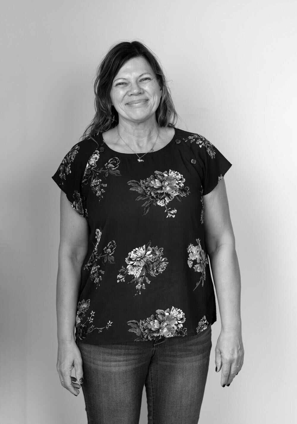 A black and white portrait of a woman in a cardigan, smiling gently with hands folded—capturing a natural moment before her transformation.