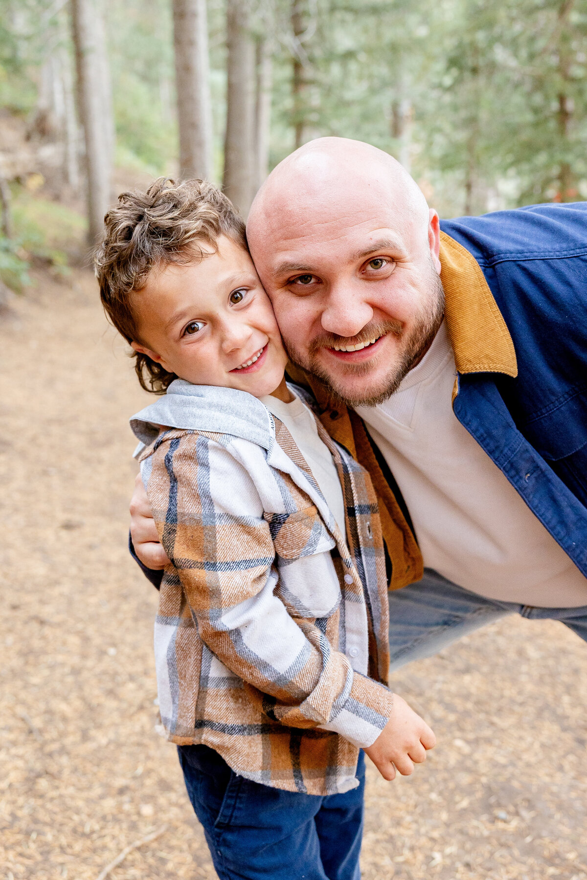 A father crouches down and embraces his young son, both smiling at the camera, surrounded by the beauty of a wooded trail.