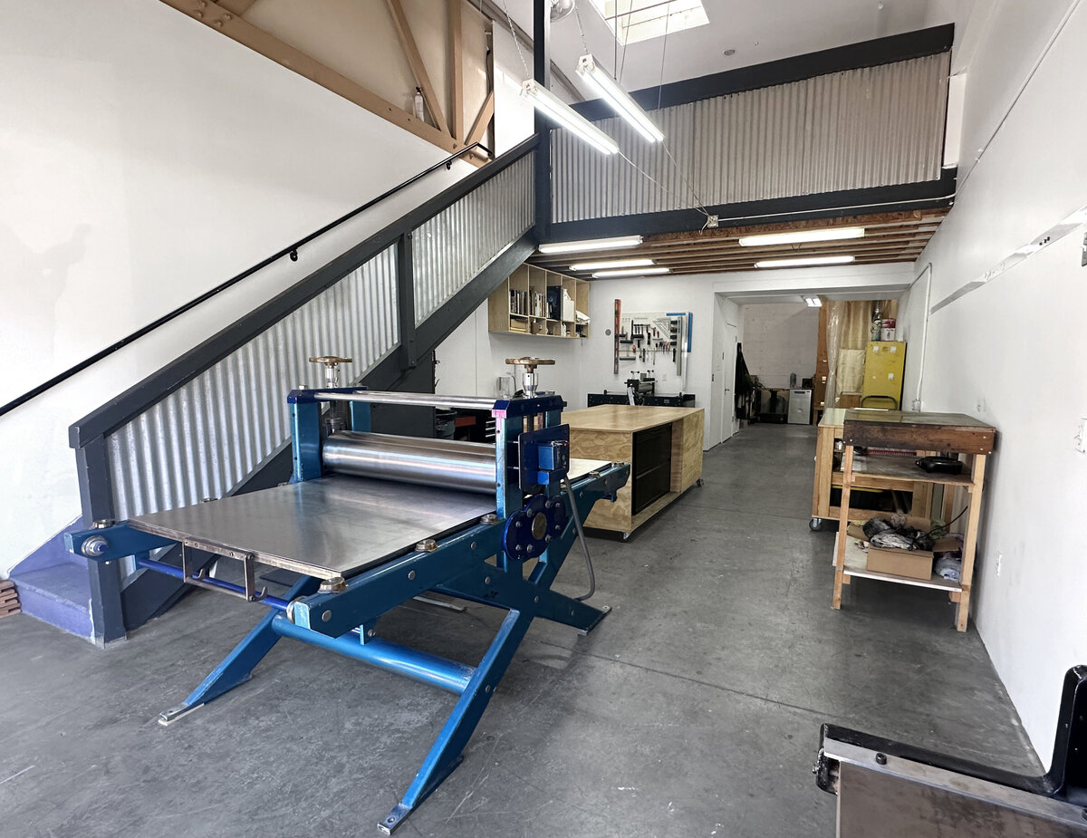 Wide-angle view of a printmaking studio showing a large blue press machine, work tables, and a neat storage area for tools and materials.