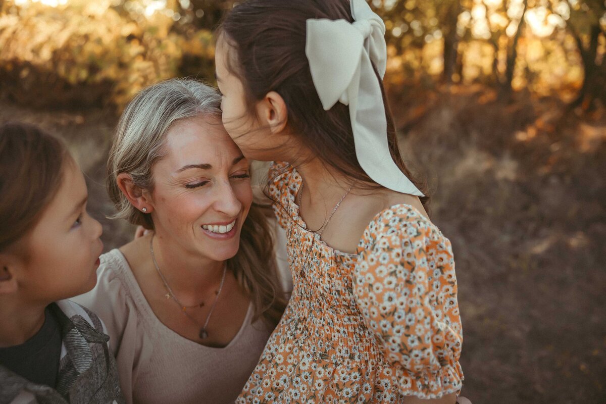 Daughter is kiss her mother on the forehead. The girl is wearing a big white bow and a ditsy flower dress.