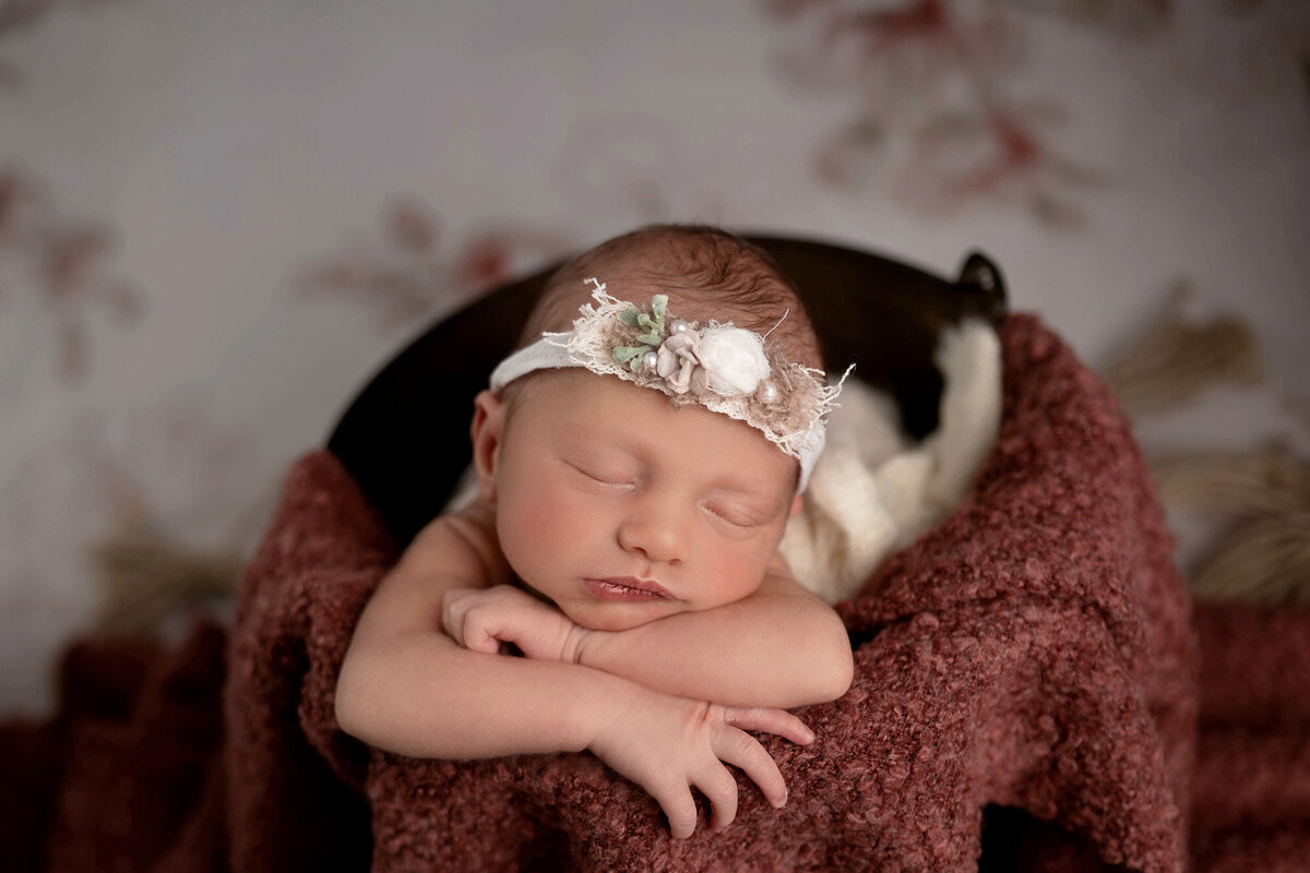 An image captured by a Pella Newborn Photographer of a baby sleeping in a white floral headband in a bucket on a red blanket