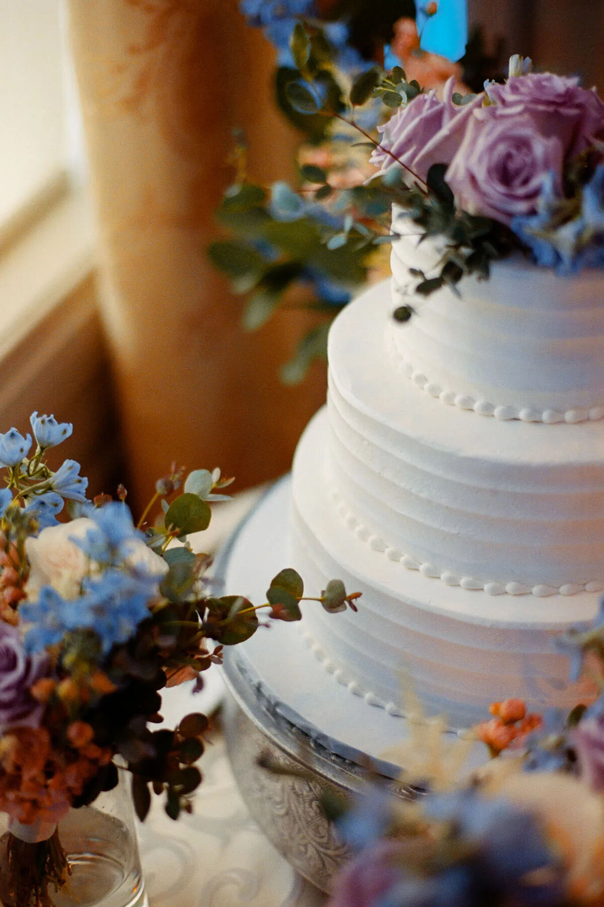 A white, three-tiered wedding cake decorated with purple roses, blue flowers, and greenery, displayed on a silver stand beside floral arrangements, beautifully captured by an NJ wedding photographer.