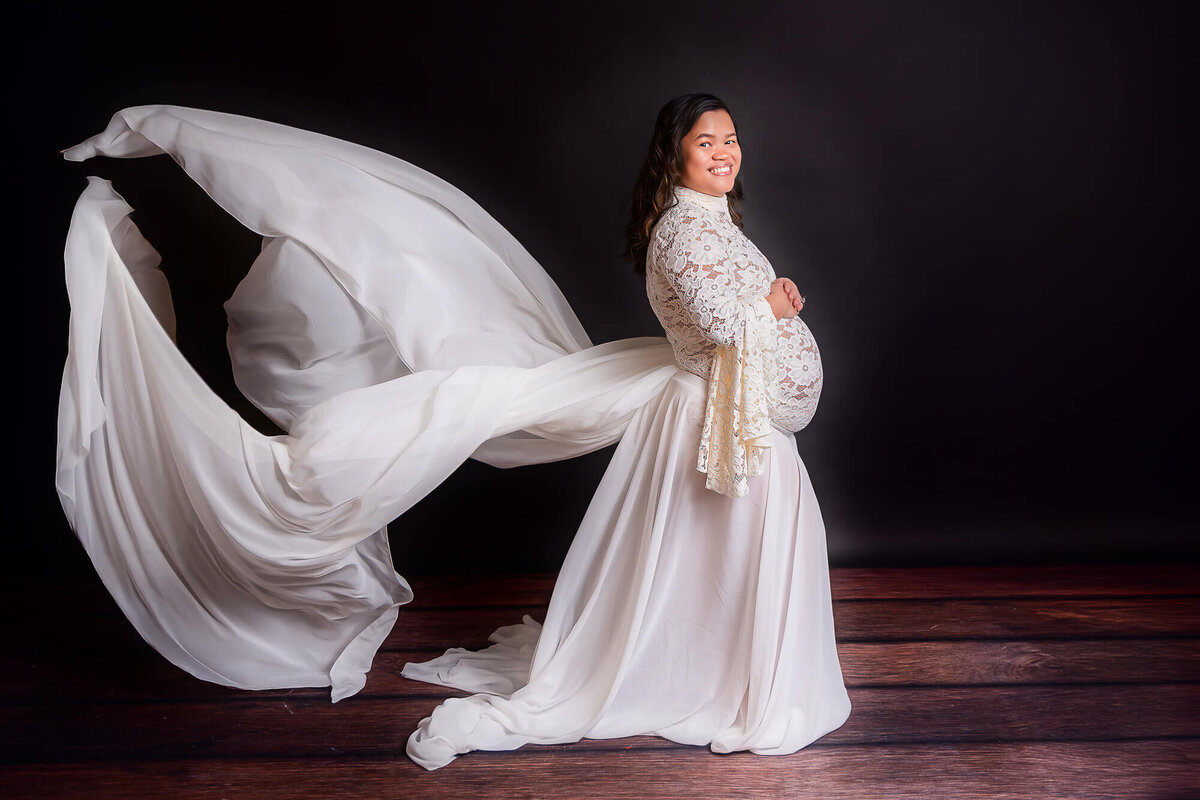 A glowing pregnant woman in a white dress stands with her arms wide open, celebrating her journey into motherhood.