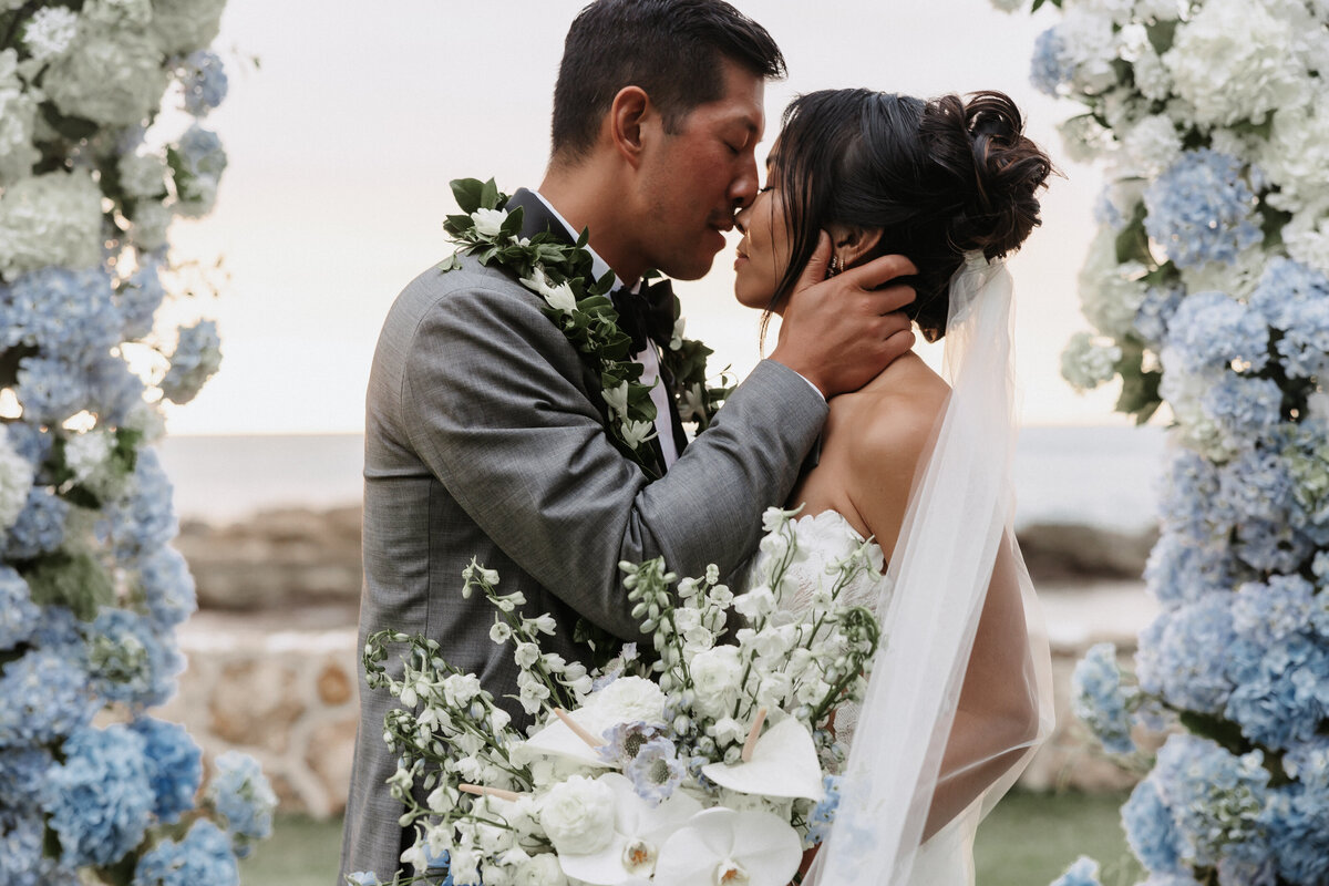 wedding couple married at lanikuhonua in ko'olina in front of blue and white bunches of hydrangeas with the ocean behind them, about to kiss.