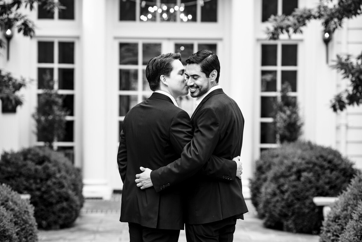 Black-and-white-image-of-a-gay-couple-with-their-arms-around-each-other-and-one-giving-the-other-a-kiss-on-the-cheek-at-The-Duke-Mansion-in-Charlotte-NC