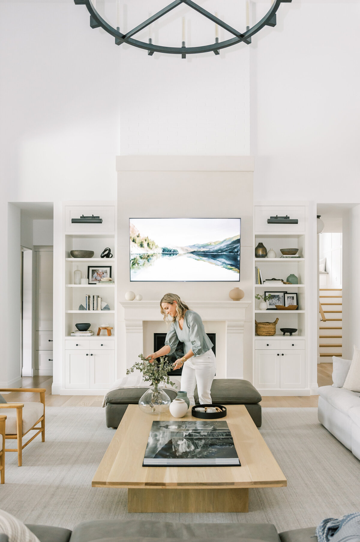 Premiere Sarnia, Ontario, Canada Interior Designer is captured arranging eucalyptus greenery in her stylish and functionally designed living room. She is looking down at the tall greenery  on an oversize coffee table with a fireplace and sytled bookshelves in the background.