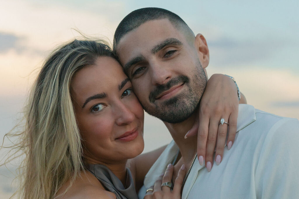 An engaged couple embraces and smiles towards the camera during sunset.