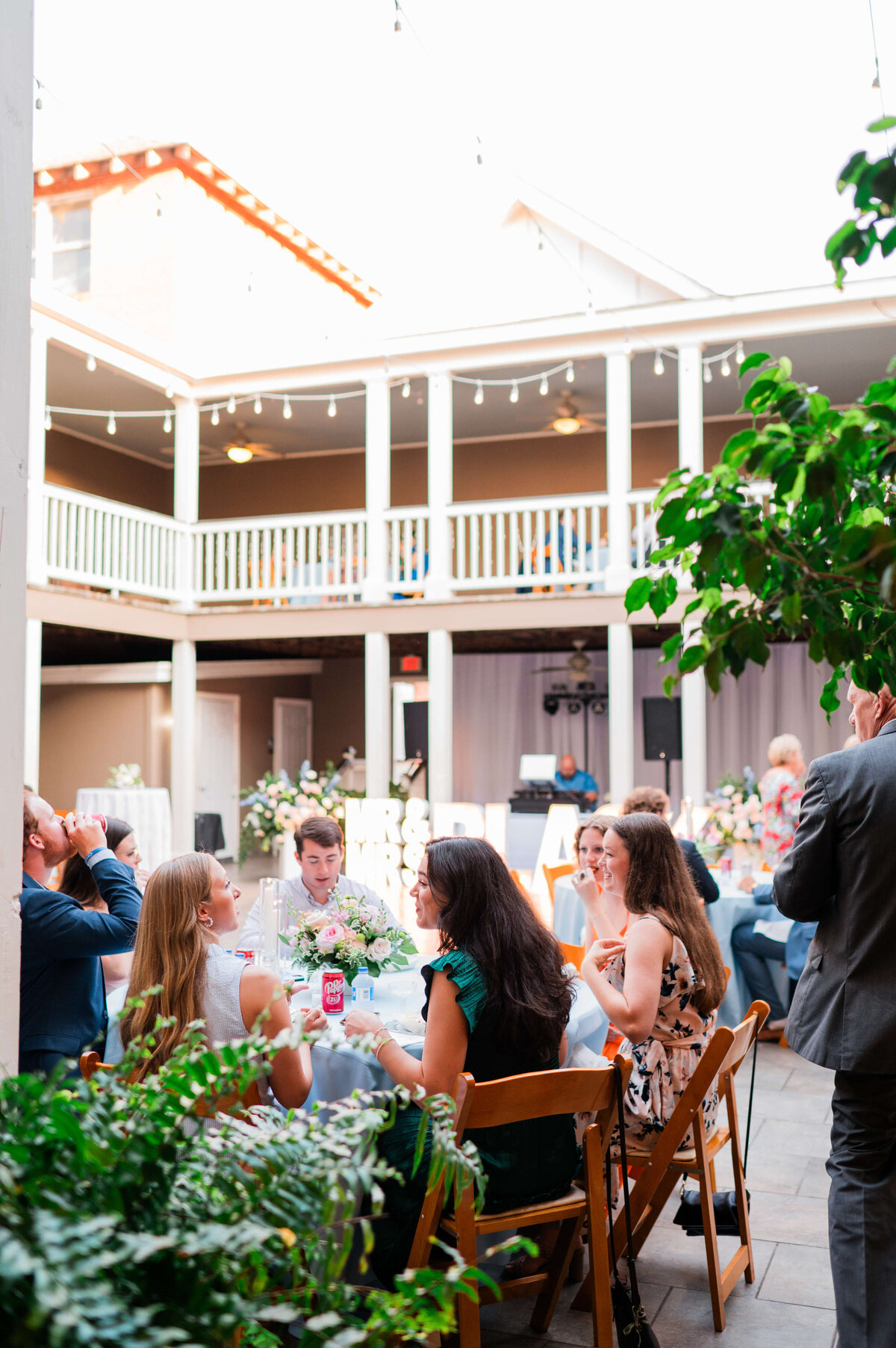A group of people sit around decorated tables at an outdoor wedding reception, surrounded by plants and string lights, with a balcony in the background.