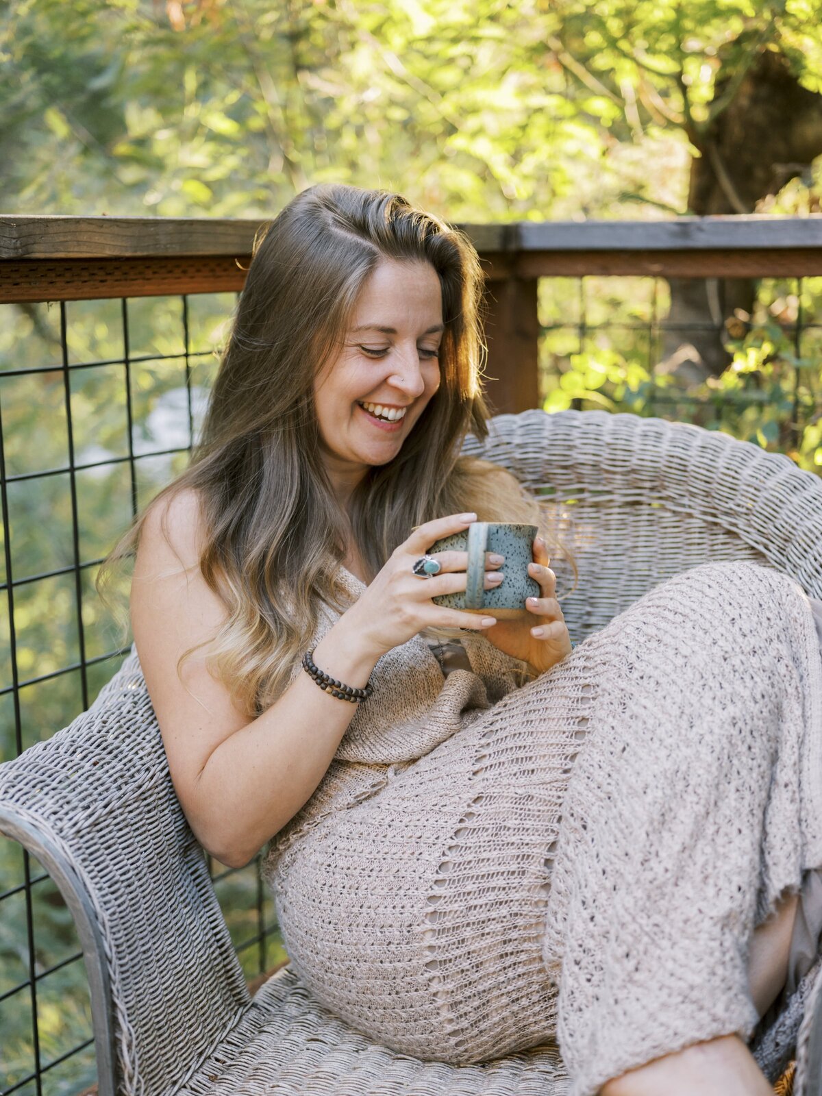 Woman sitting in a wicker chair holding a coffee mug outside on her porch and smiling away from the camera