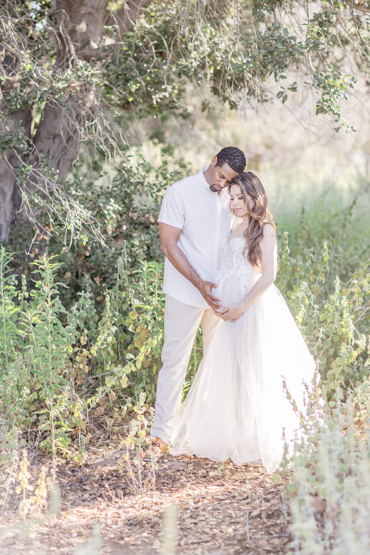 A couple stands in a sunlit forest clearing, gently embracing. The woman is wearing a flowing white dress, while the man is in a white shirt and light pants. They are surrounded by lush greenery, with sunlight filtering through the trees.