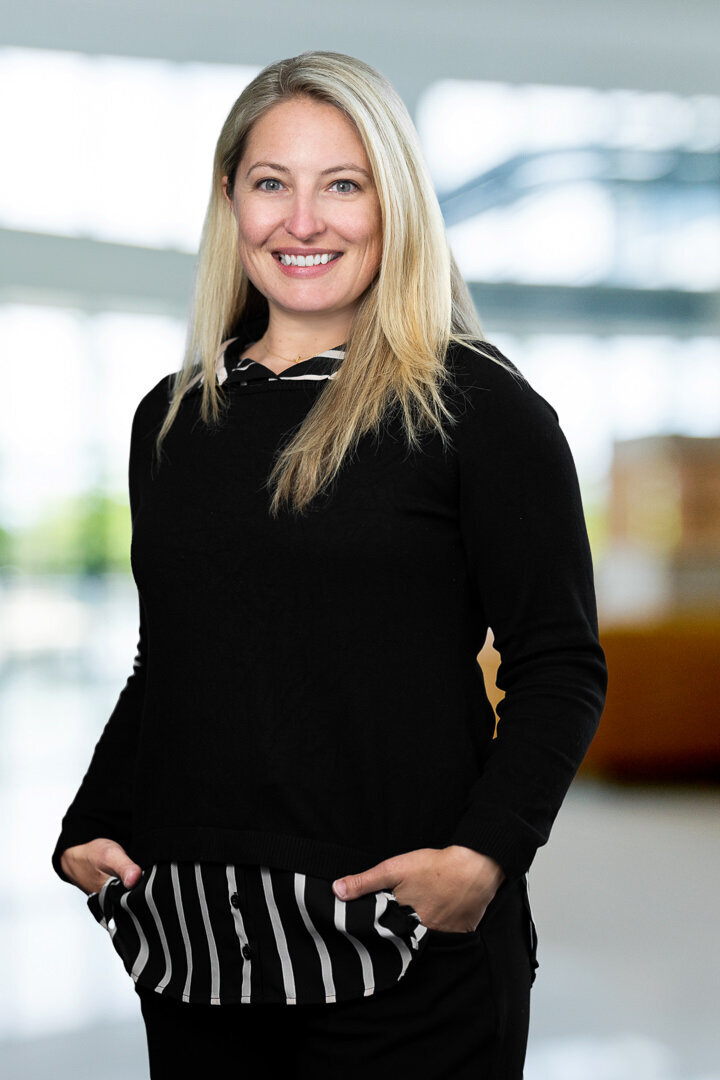 Professional headshot of smiling blonde woman in black sweater, approachable business portrait with blurred office background by Alex Kaplan Photo.