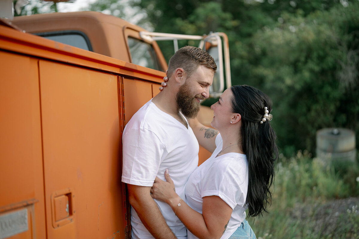 Kali and Joe hugging beside a rusty orange vintage truck during their engagement session on private property near Detroit.