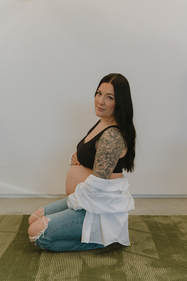 Kneeling on the ground, a pregnant woman gently holds her belly while softly smiling at the camera.