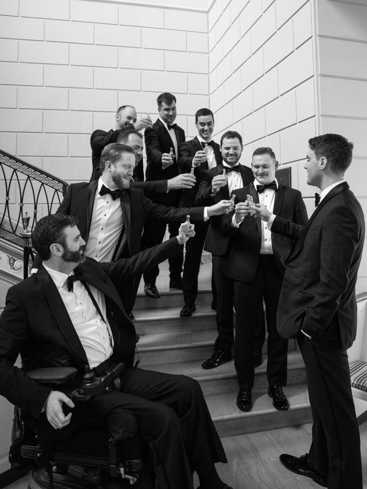 A group of men in tuxedos celebrate cheerfully on a staircase, holding glasses in a toast. One man is in a wheelchair. The atmosphere is joyful and festive, suggesting a formal event or celebration. Black and white photo.