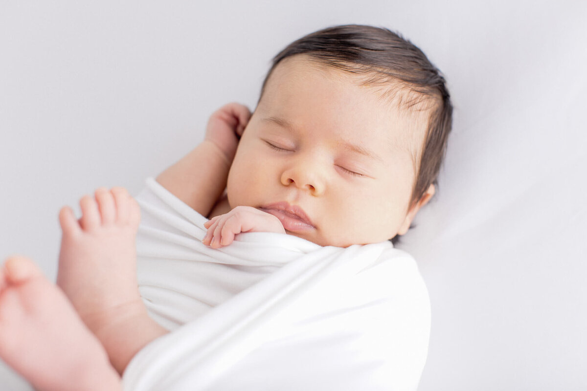 A sleeping baby wrapped in a white blanket lies peacefully on a white surface, with one hand near the face and feet slightly exposed. The baby has dark hair and a calm expression.