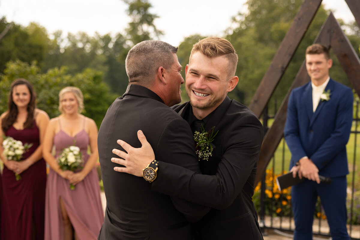 Groom embraces his new father-in-law as his bride walks down the aisle on their wedding day. Photographed by Aaron Aldhizer