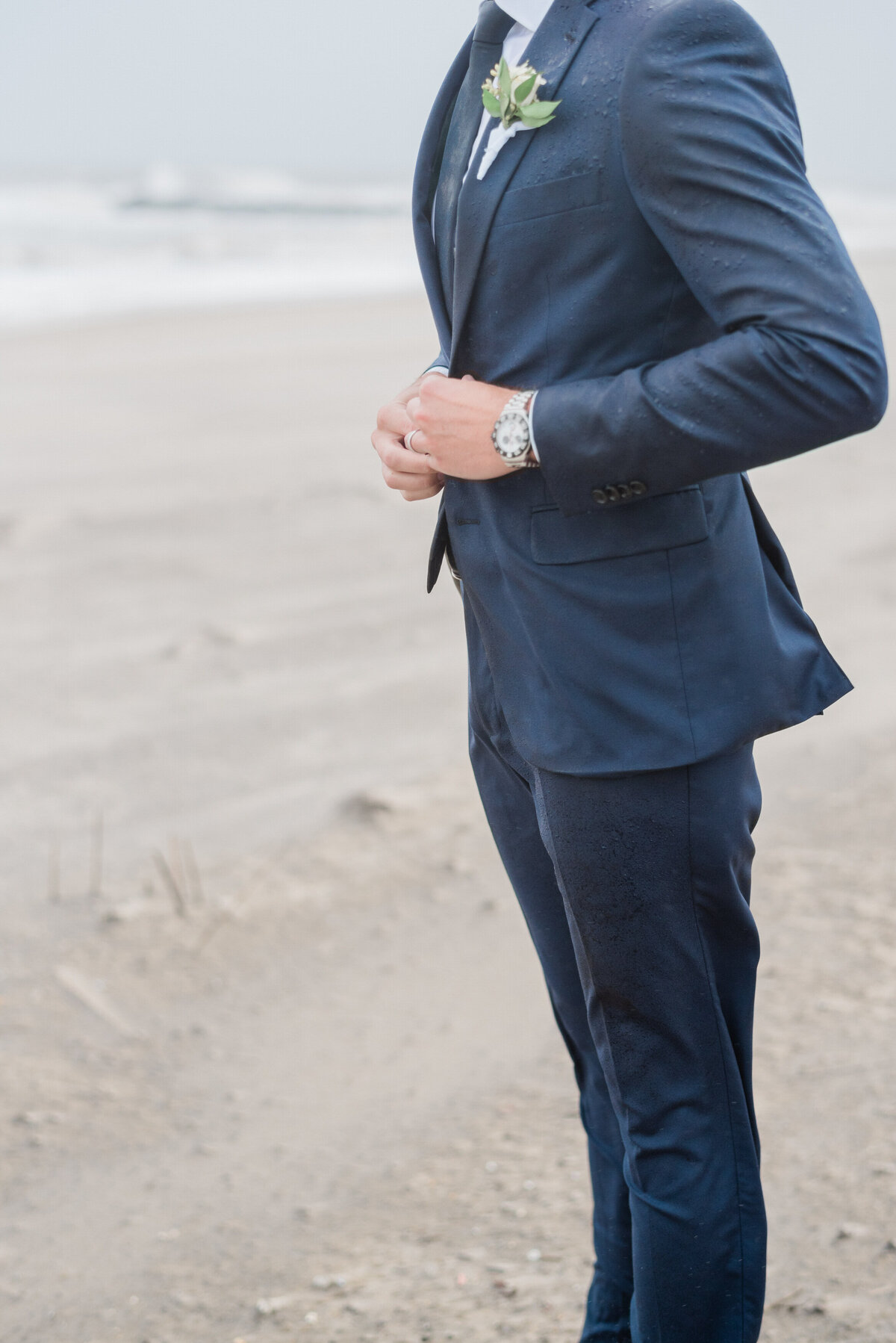 the groom buttoning his navy blue suit jacket, with the ocean in the background