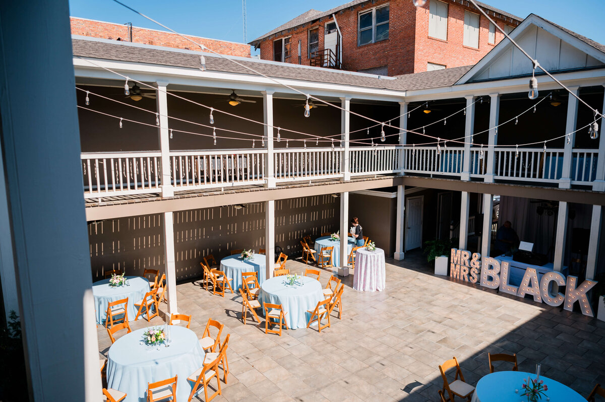 An outdoor courtyard set up for an event with round tables covered in light blue cloths, wooden chairs, string lights overhead, and large MR and MRS letters near a wall.