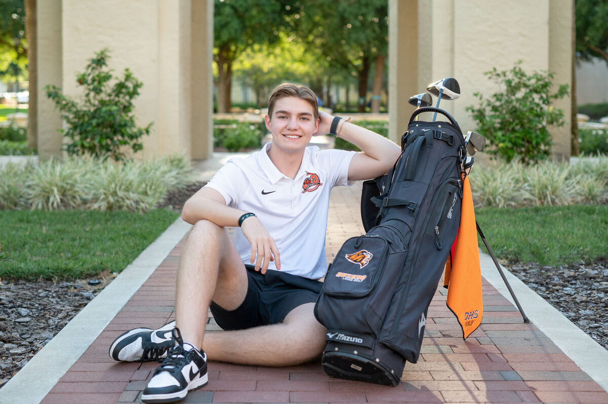 High school senior boy sitting on floor posing next to golf bag.