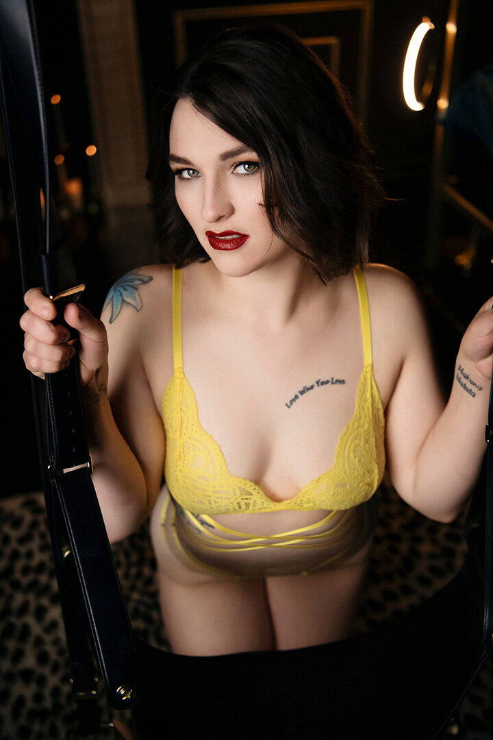 A woman with short dark hair and red lips, wearing a yellow lace bra, holds black straps. She gazes upward with confidence in a dimly lit room.
