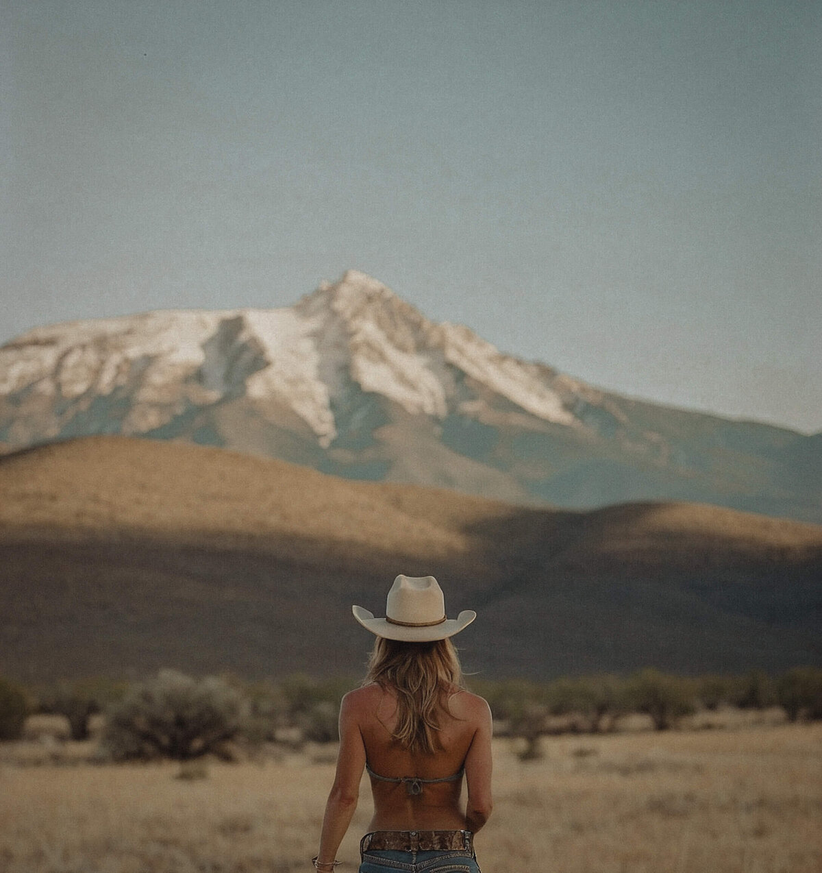 A woman wearing a cowboy hat stands in an open field, facing a distant snow-capped mountain under a clear sky—a scene as thoughtfully crafted as Sweet Pea Ink Creative’s brand design.