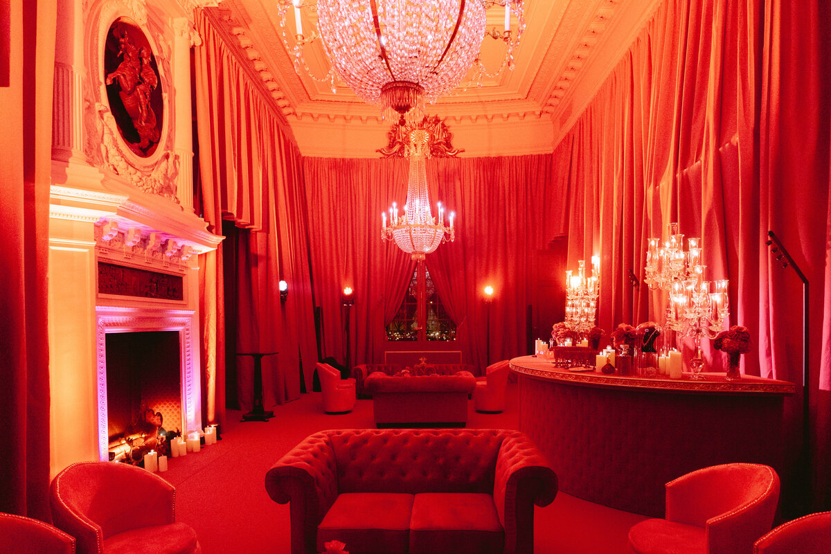 Lavishly decorated room illuminated by red lighting, featuring a large chandelier hanging from the ornate ceiling, plush couches, a grand fireplace with lit candles, and a well-stocked bar. The elegant setting exudes a warm and luxurious ambiance.