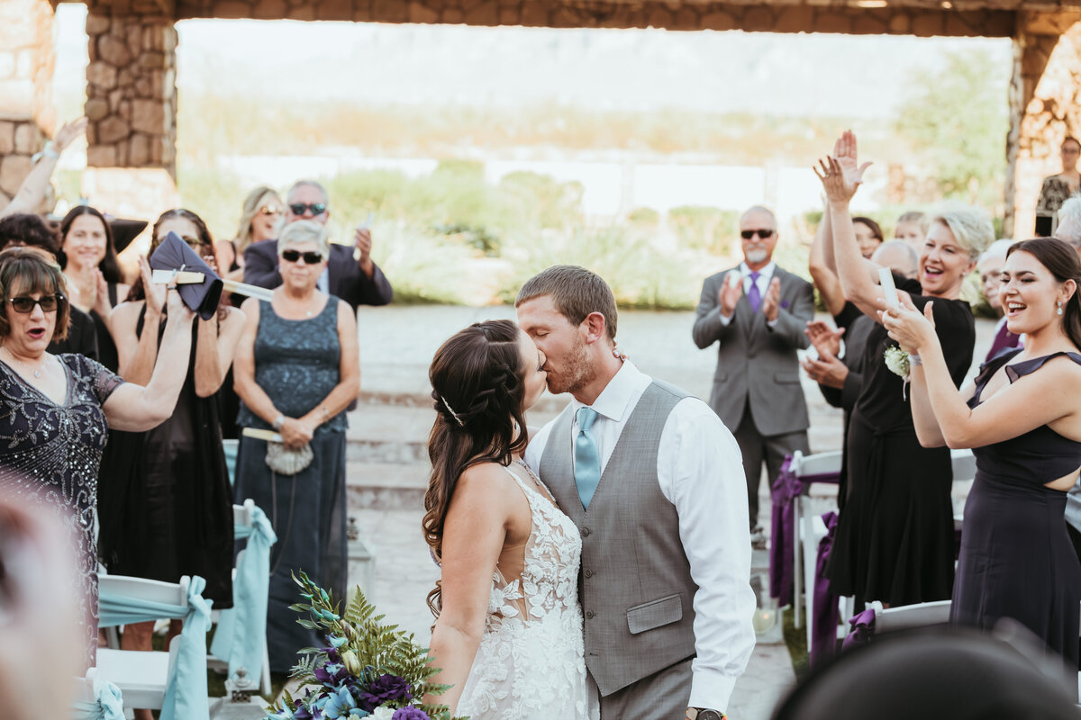 A joyful bride and groom kiss during their wedding ceremony, surrounded by cheering guests. The attendees, varying in age and dressed in formal attire, express happiness and excitement; some are clapping, and others are capturing the moment on their phones and cameras. The outdoor setting is adorned with natural greenery and rustic wooden elements, creating a festive and heartwarming atmosphere.