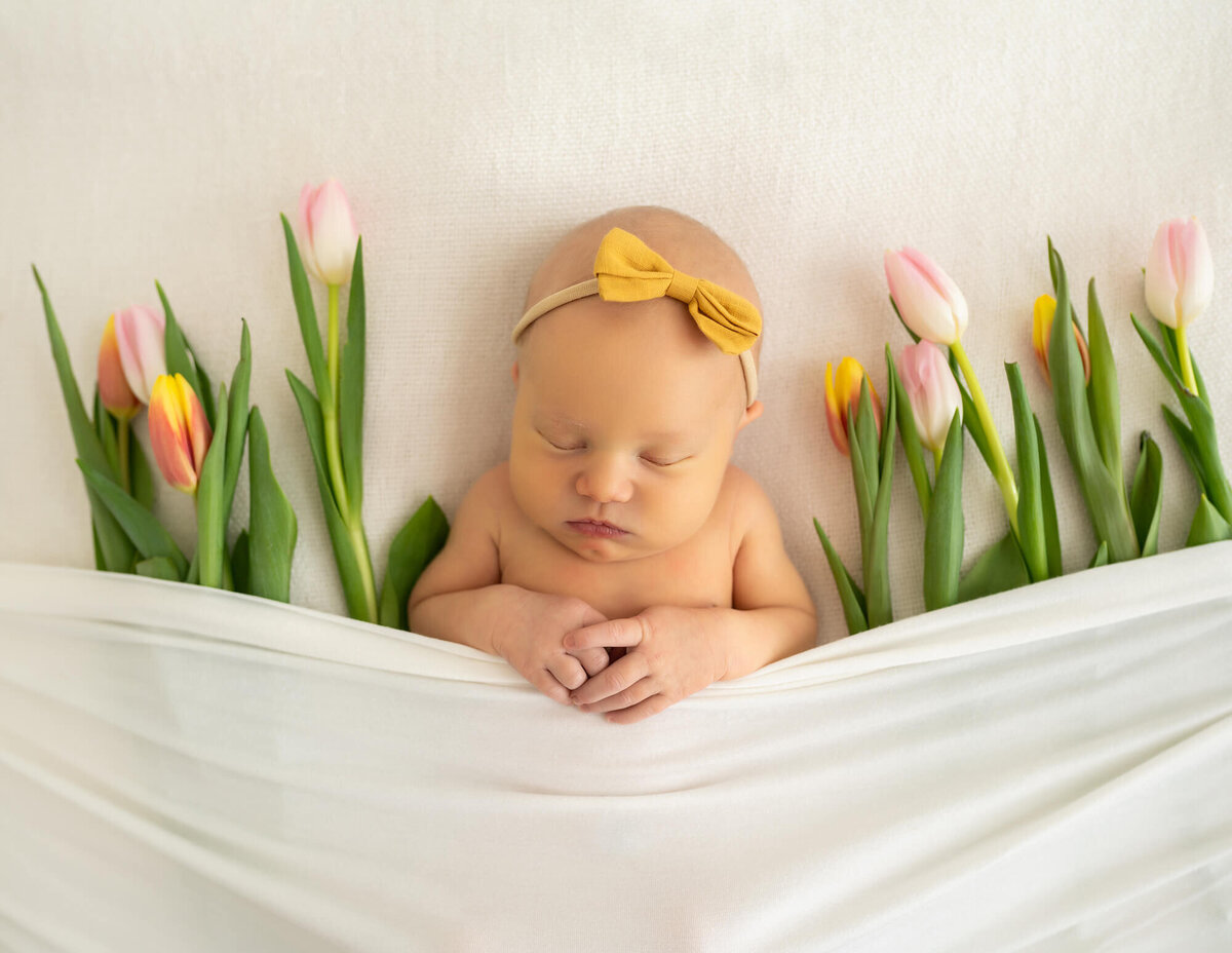 A newborn baby girl with a yellow headband lays under a white blanket surrounded by tulips