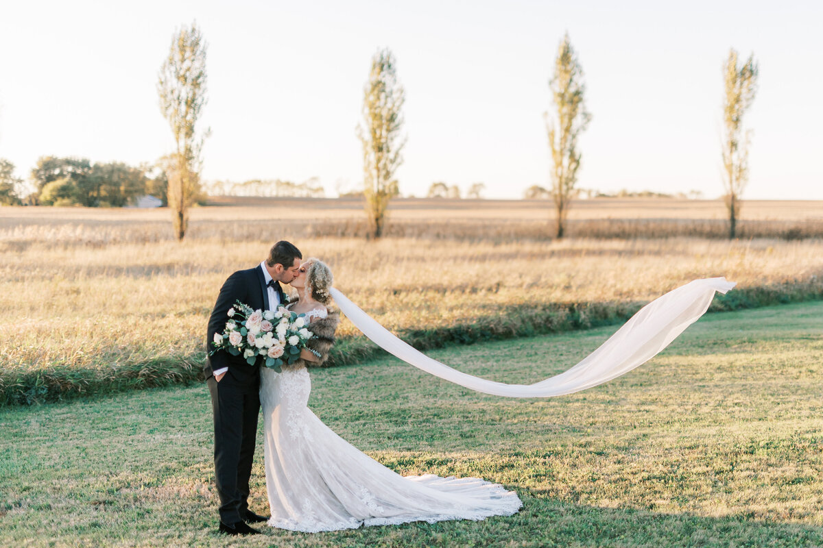 Bride and groom kiss while holding bouquet, as wedding veil blows in the breeze