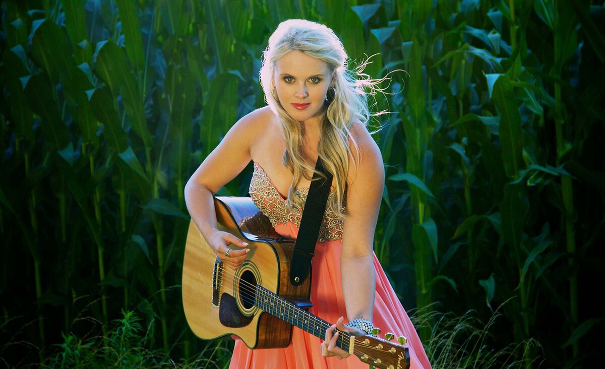 Female musician portrait Krista Earle wearing pink dress playing guitar against corn field background