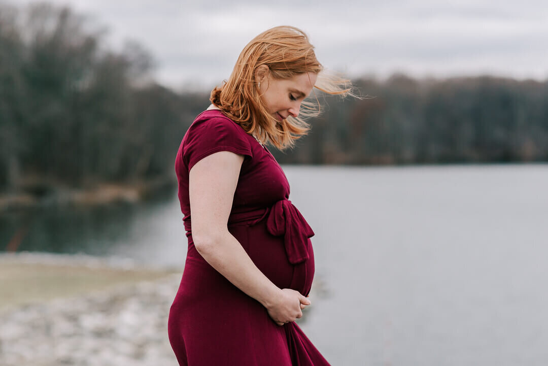 A red-haired woman looking down at her beautiful growing belly, against a gorgeous cloudy lake backdrop