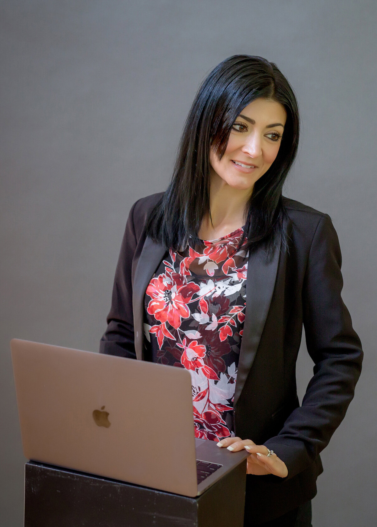 A female professional speaker wearing a floral shirt & black blazer standing at a podium with a laptop, presenting to an audience, being photographed by a South Shore MA branding photographer