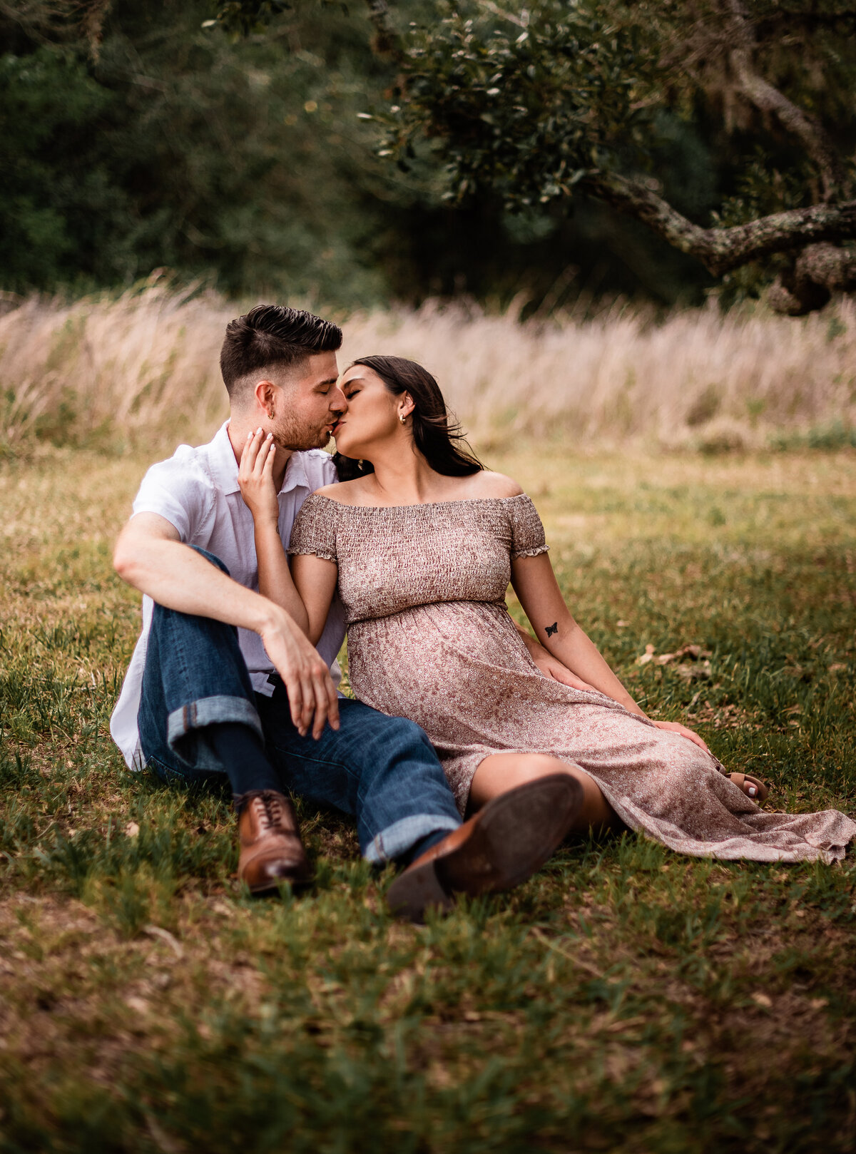 A couple sits near a field of long grass and cuddles close while they kiss.