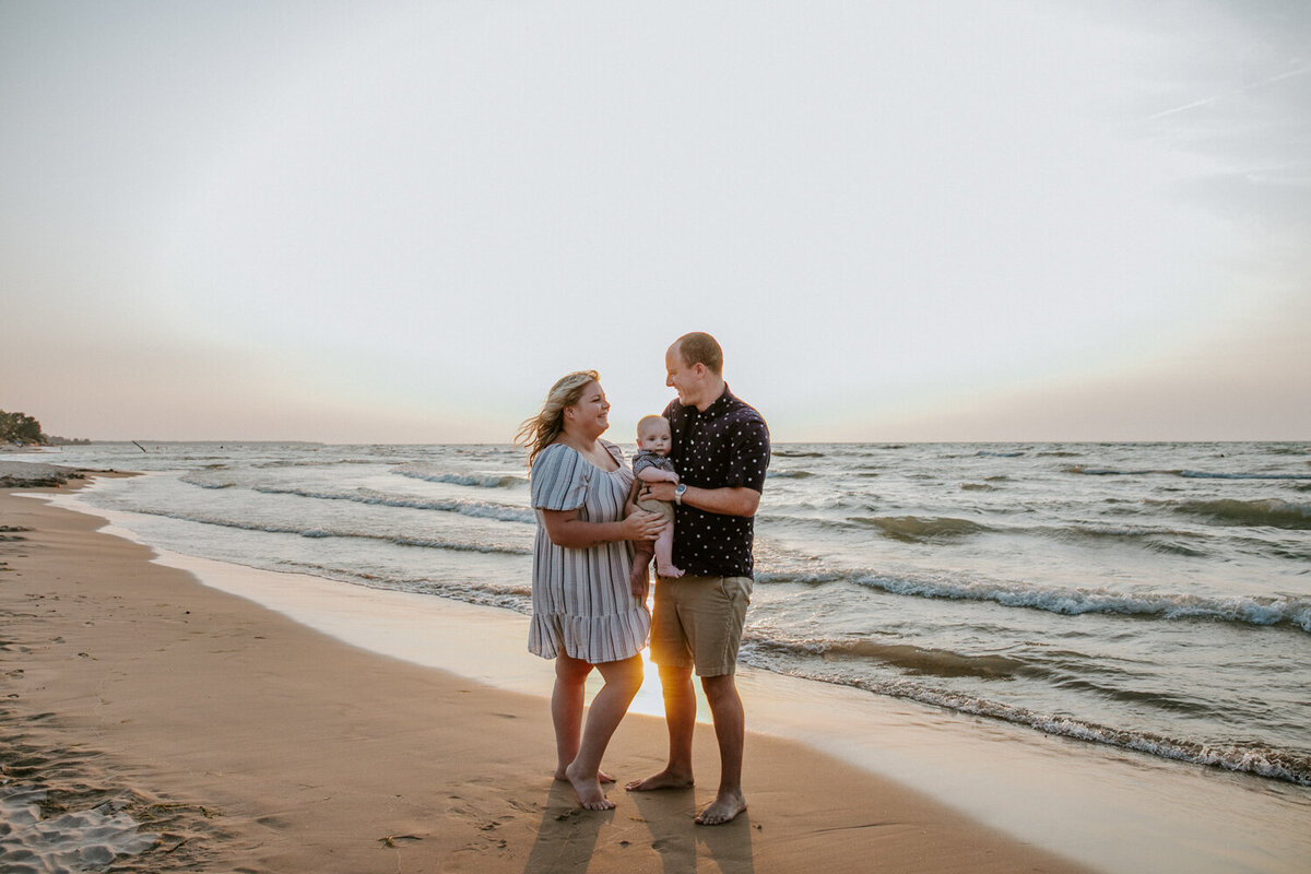 Mom, Dad and infant stand at the Grand Bend beach shoreline for top family photographer. Mom and Dad are  smiling at each other, holding the baby between them. The baby is looking at the camera. The sun is setting behind them.