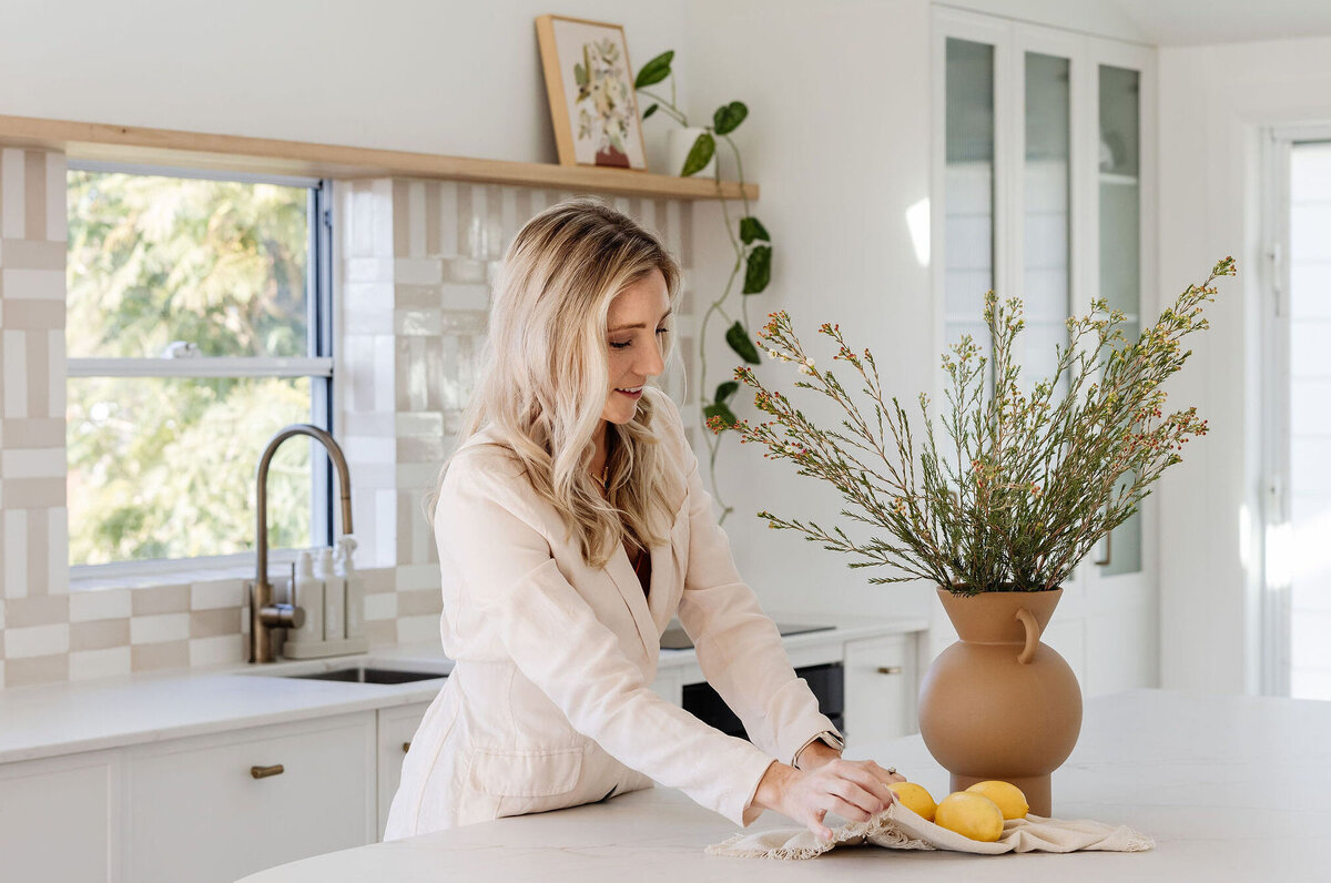 In a beautifully curated space reflecting bespoke interior design in Newcastle NSW, a woman carefully arranges lemons and a vase on the pristine white kitchen counter, accented by lush plants adorning the background.