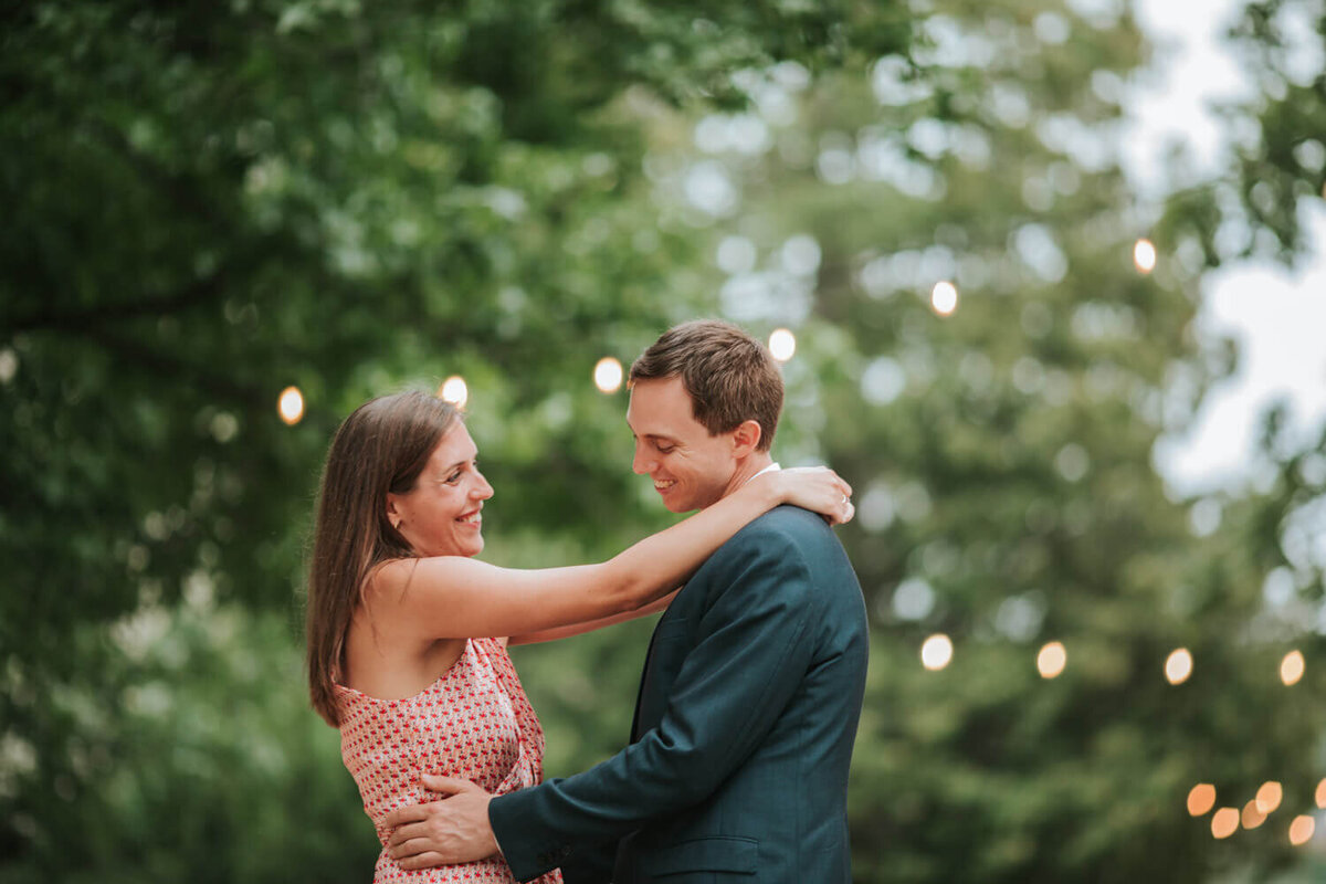 A couple shares a joyful embrace under string lights, surrounded by lush greenery