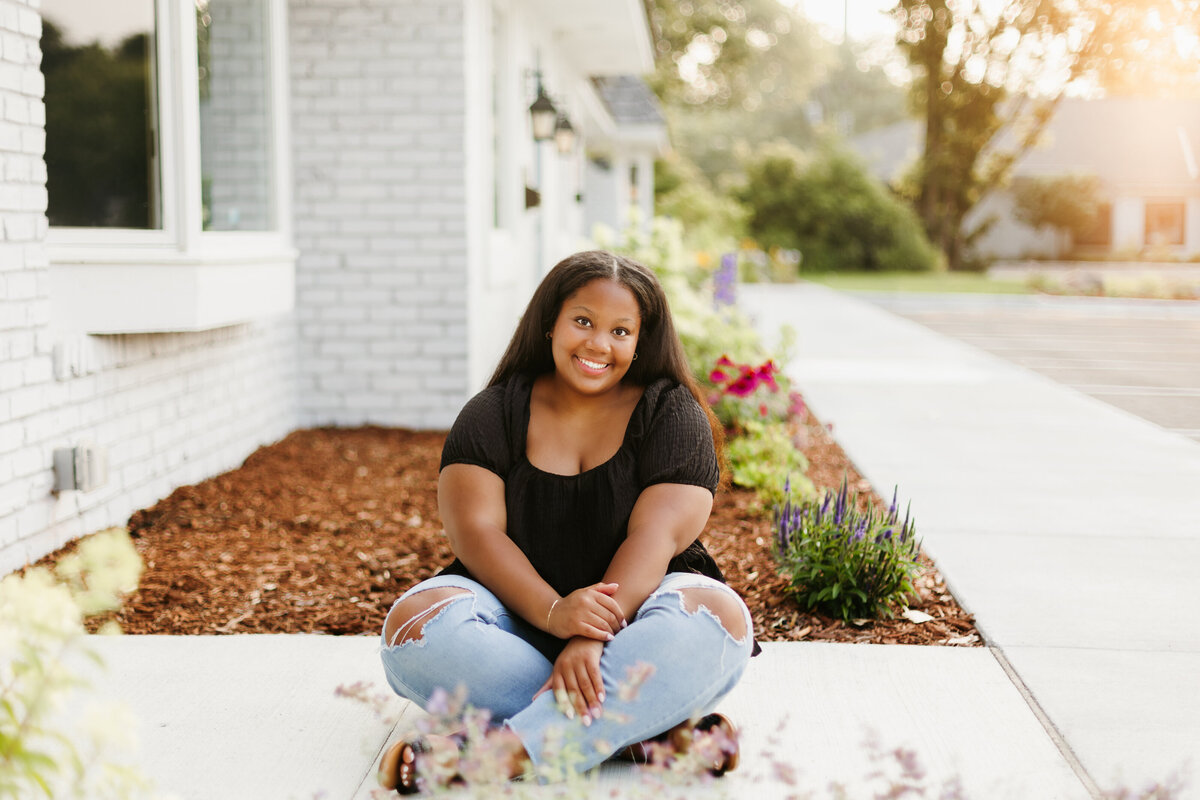 Senior girl in a black top and jeans sitting cross legged on a sidewalk