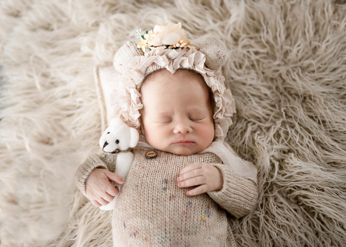 A baby sleeps on a fur blanket while cuddling a tiny bear with a floral bonnet