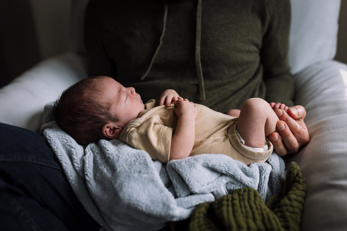 newborn in tan onesie laying on light blue and olive green blankets in dad's lap with dad holding his feet
