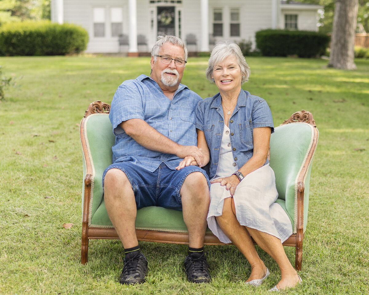 Grandparents on sitting on the vintage green couch in the grass with their family home in the background, smiling at the camera.  Grandpa is wearing a light blue and white stripped short sleeve button up shirt, blue jean shorts, and has white short hair and goatee.  Grandma is wearing a white button up dress with a jean jacket over it, with short white hair.  
