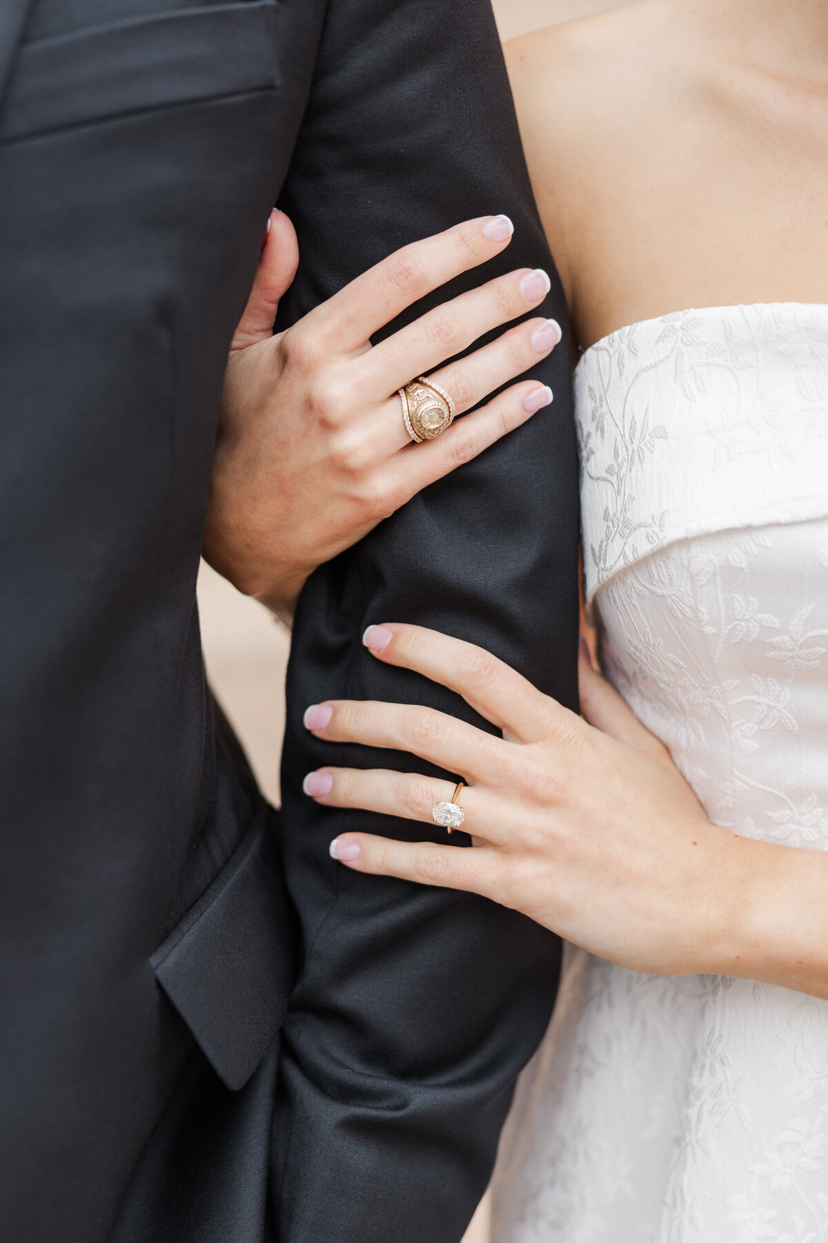 A detail shot of a DFW couple during their engagement session, highlighting their intertwined arms and stunning rings. The bride-to-be wears an embroidered white dress and a radiant oval-cut engagement ring, while the groom sports a class ring and black suit. A timeless image full of love and elegance.